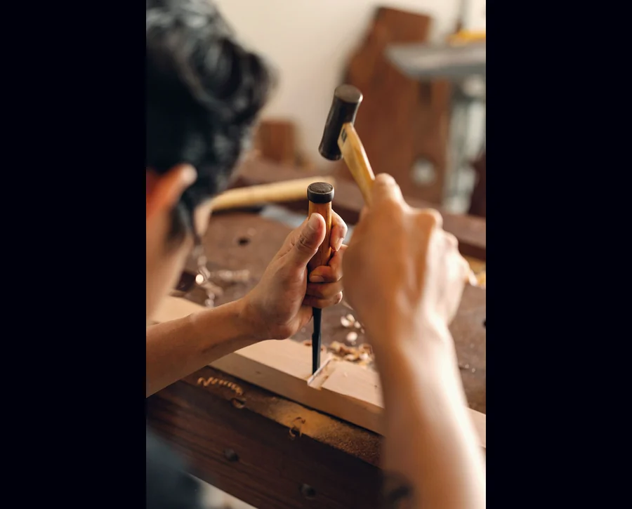 A joiner's hands guiding a chisel and mallet across a length of timber at the bench.