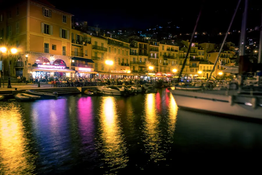 Harbour-front restaurants lit at night, their colours reflected in the water among moored boats.