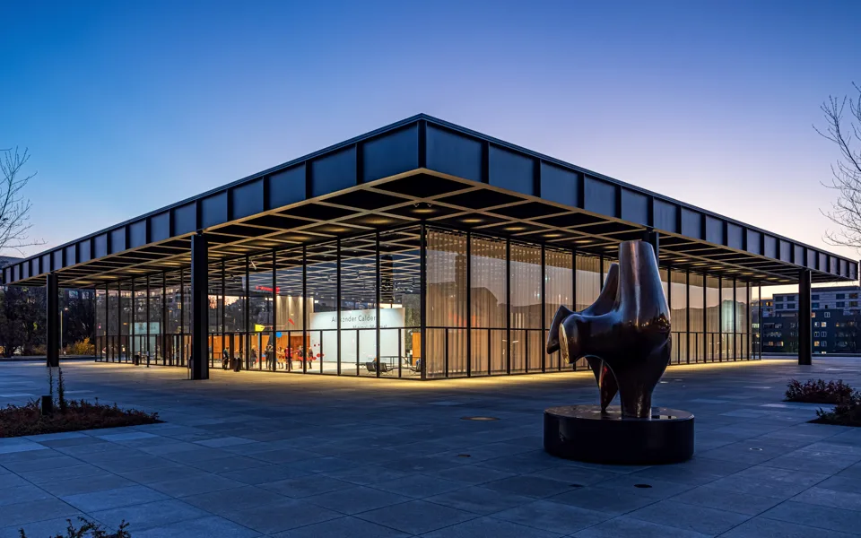 A steel and glass pavilion at dusk, its interior lit warmly beneath a deep blue sky.