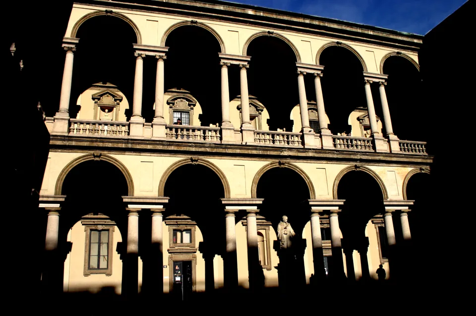 Two storeys of stone arcades in a Milanese palazzo courtyard, in hard sunlight and deep shadow.