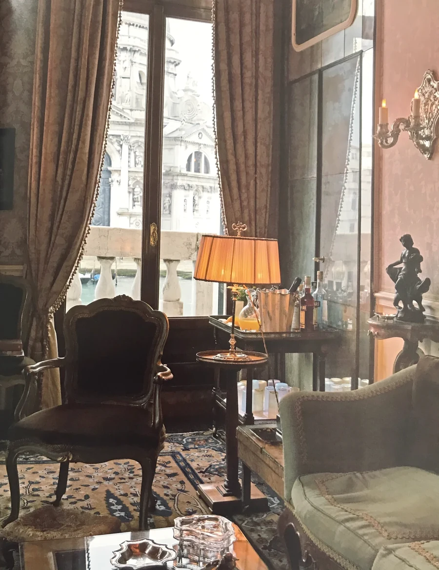 A corner of a salon at dusk, a silk-shaded lamp lit beside tall windows opening onto a baroque façade across the water.