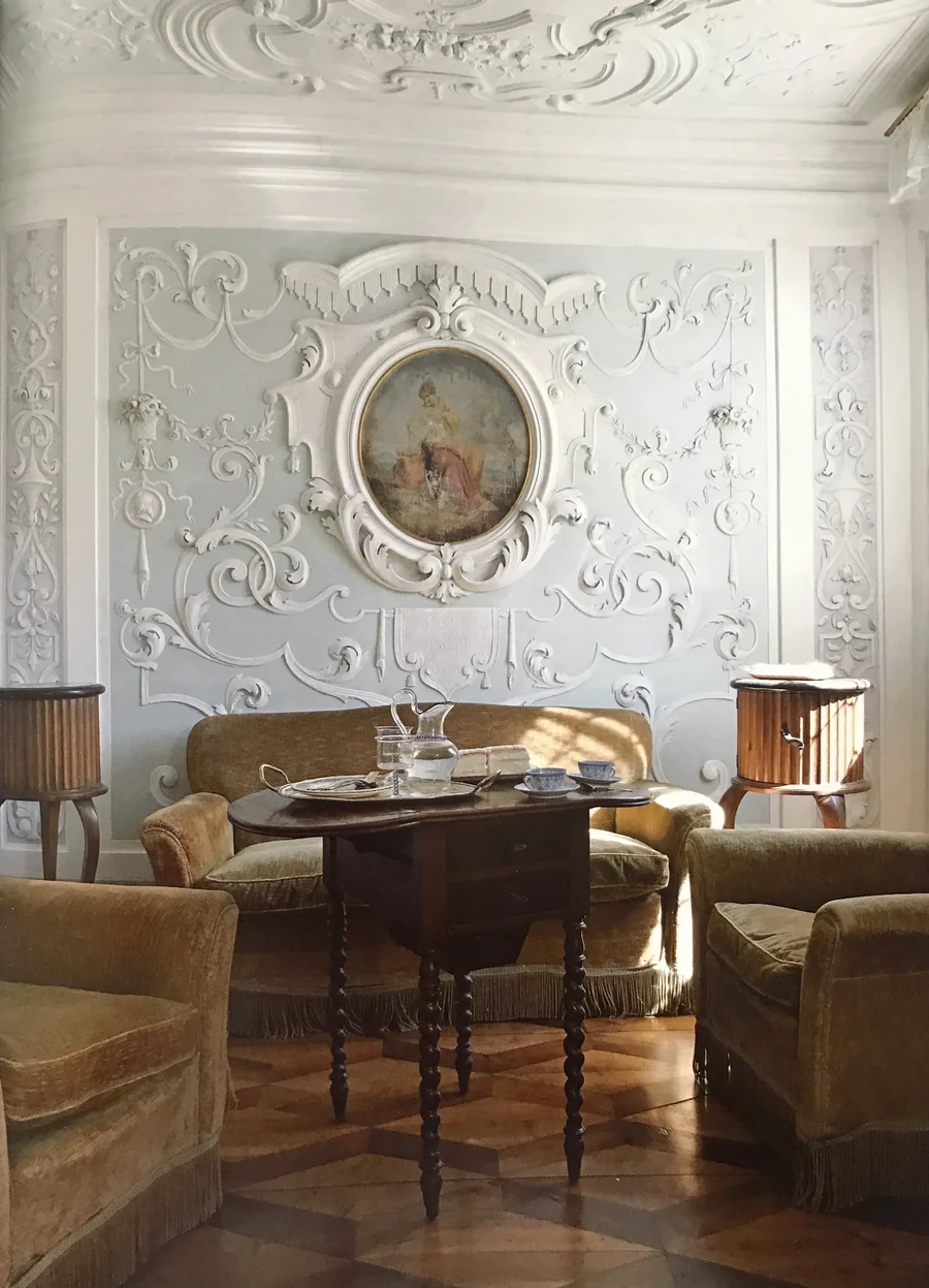 A stuccoed salon in low winter light, a tray of coffee set on a small walnut table between velvet armchairs.