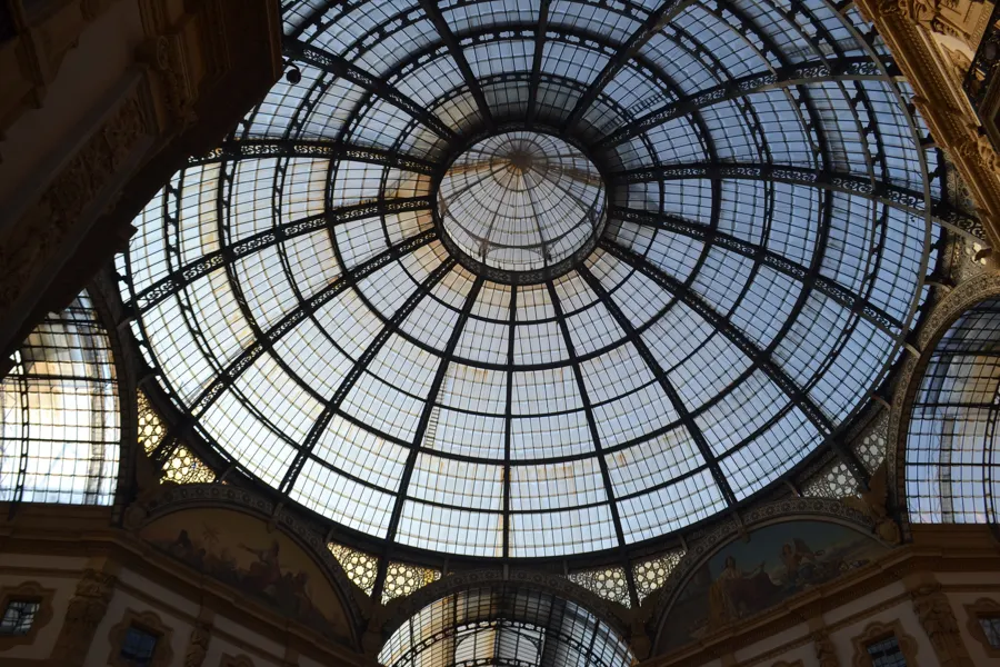 The iron and glass dome of a Milanese gallery seen from below, with painted lunettes.