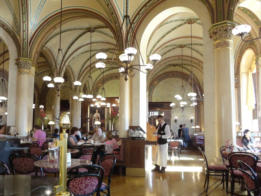 Guests at marble tables beneath the vaulted ceilings and lamps of a Viennese coffee house, a waiter standing by.